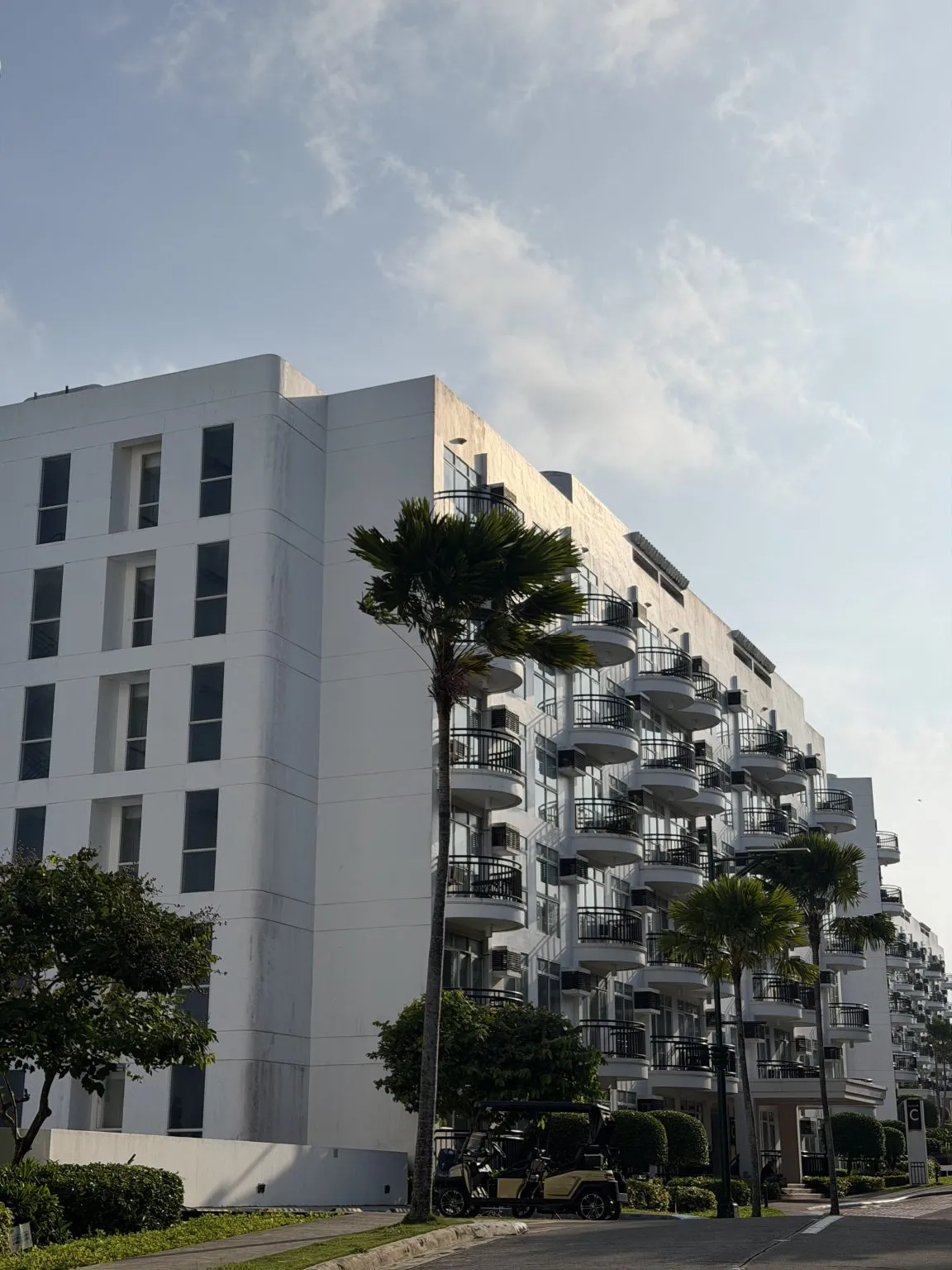 A white multi-storey residential building with rows of rounded balconies behind palm trees.